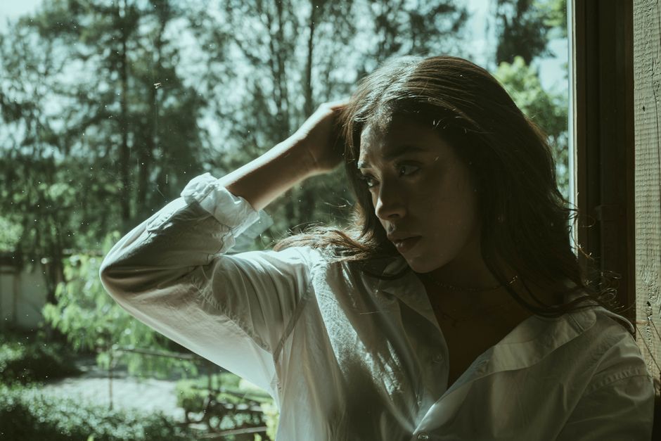 A thoughtful woman stands by a sunlit window, embracing natural light in a serene setting.