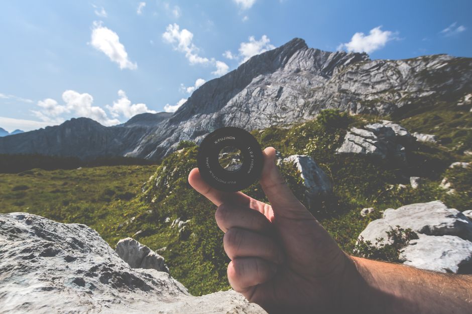 A hand holding a camera lens against a scenic mountain backdrop under a clear sky.